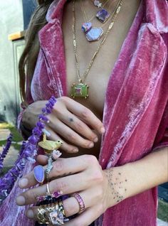 a woman in a pink robe holding several rings and bracelets on her hand with purple flowers behind her