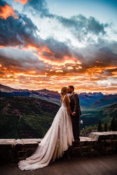 a bride and groom standing on the edge of a cliff at sunset with mountains in the background