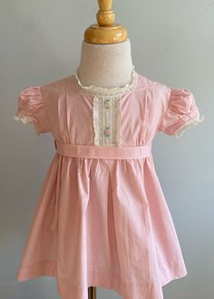 "This pretty dress was made from a pink broadcloth and the label is \"Nathan Krauskoff\". The construction of the bodices is a little different in that pleats are used. The front bodice has two pleats either side of center. These are sewn down from shoulder for about 2\", then released down to waist. At center front is a pretty lace trim with openwork and pastel floral embroidery. Directly above is the squared neckline with a white lace frill. Below waist, there is a center inverted pleat flanke Classic Ruffled Dress With Fitted Bodice, Classic Daywear Dress With Smocked Bodice, Classic Empire Waist Dress For Daywear, Fitted Pink Dress With Pleated Bodice, Pink Pleated Dress With Fitted Bodice, Pink Fitted Dress With Pleated Bodice, Classic Vintage Dress With Fitted Bodice For Spring, Classic Vintage Dress With Ruffles For Spring, Classic Short Sleeve Dress For Dress-up