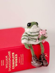 a small stuffed frog sitting on top of a red book holding a pink object in it's hand