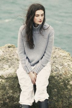 a woman sitting on top of a rock next to the ocean with her hands in her pockets