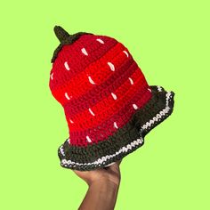 a hand holding up a knitted hat on top of a green background in the shape of a watermelon