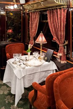 a dining car on a train with red chairs and tables set for two, along with a chandelier hanging from the ceiling