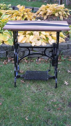 an old sewing table is sitting in the grass next to some bushes and flowers with yellow leaves on it