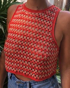 a woman wearing a red crocheted crop top standing next to a plant and looking at the camera