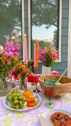 a table topped with plates of food next to a bowl of fruit and flowers in front of a window