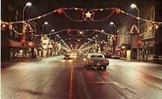 cars driving down an empty city street with christmas lights strung across the street and buildings on both sides