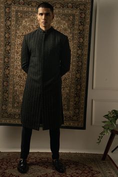 a man standing in front of a rug wearing a black coat and dress shirt with pleated details