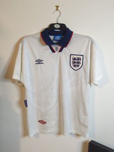 This classic England home shirt from 1993-95 is a must-have for any football fan. With the iconic Umbro branding and short sleeves, it's the perfect way to show your support for the national team. Made for adults in size medium, this shirt is suitable for both casual wear and for display as a sports memorabilia item. The white design with the red and navy accents make it a timeless piece that will never go out of style. Whether you are a diehard supporter of the Three Lions or just appreciate th White Team Logo Shirt For Sports Events, White Shirt With Team Logo For Sports Events, White Shirt With Team Logo For Sports Season, White Team Logo Shirt For Fan Merchandise, White Shirt With Team Logo For Fans, White Shirt With Team Logo For Fan Merchandise, White Shirt With Team Logo For Team Spirit, White Team Spirit Shirt With Logo, White Shirt With Team Logo