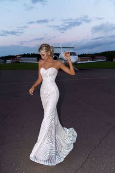 a woman in a white dress is walking on the tarmac with an airplane behind her