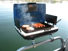 an outdoor grill with hot dogs cooking on it's side and water in the background