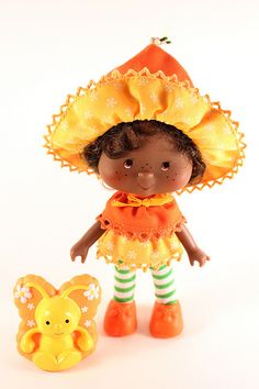 a doll with an orange dress and yellow hat next to a small toy rabbit on a white background