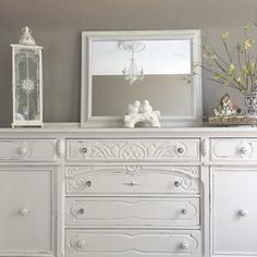 a white dresser with two mirrors on top of it and flowers in vases next to it