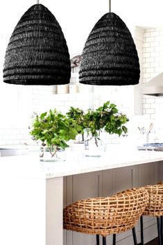 two hanging lights above a kitchen island with plants in it and wicker chairs at the counter