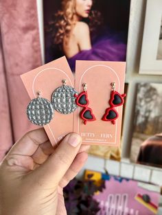 a person holding up two pairs of earrings in front of a pink card with an image of a woman on it