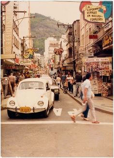 an old photo of people walking on the sidewalk in front of cars and buildings with signs above them