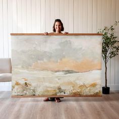 a woman standing behind a large painting on a wooden frame in front of a white wall