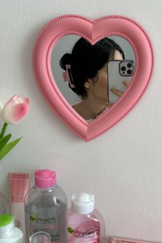 a pink heart shaped mirror with a woman's face reflected in it next to cosmetics and flowers