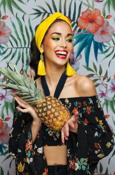 a woman wearing a yellow headband holding a pineapple in front of a floral wall