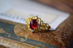 Embrace your inner royalty with the Regal Filigree Ring. Featuring a deep scarlet oval gemstone set within an intricate gold filigree band adorned with sparkling crystals, this ring exudes vintage elegance and timeless sophistication. The ornate design brings a touch of classic charm to any look, making it the perfect accessory for special occasions or adding a regal flair to your everyday style. Details Crimson Regal Filigree Ring in Red Gold pleated Adjustable opening Missy Jewelry Dragon Breath Collection Exquisite Gold Ruby Gemstone Ring, Gold Ruby Rings With Intricate Design, Elegant Jeweled Wedding Rings, Exquisite Gold Ruby Wedding Ring, Exquisite Gold Ruby Ring For Wedding, Victorian Red Oval Filigree Ring, Ornate Gold Ruby Ring, Elegant Red Crystal Gemstone Ring, Elegant Formal Jeweled Crystal Ring