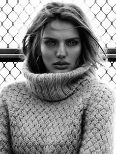 a woman wearing a cowl neck sweater in front of a chain link fence and looking at the camera