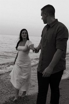 a man and woman holding hands walking on the beach next to the ocean in black and white