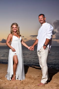 a man and woman holding hands while standing on the beach near the ocean at sunset