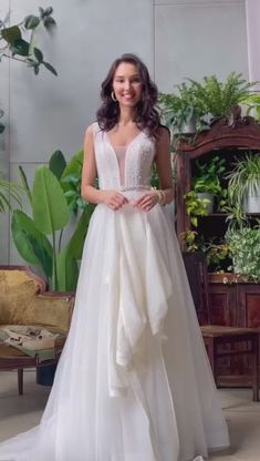 a woman in a white wedding dress standing next to a potted plant and smiling at the camera