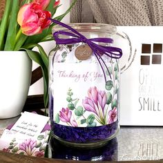 a glass jar filled with purple flowers on top of a table next to a card