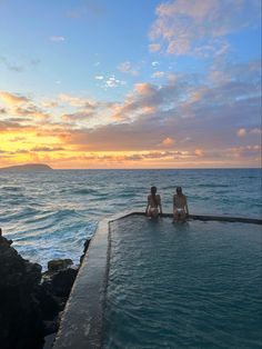 two people sitting on the edge of a swimming pool in front of an ocean at sunset