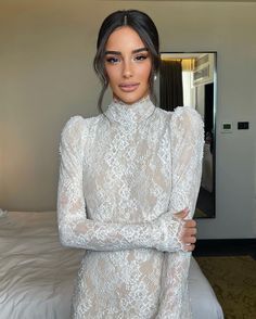 a woman standing in front of a bed with her arms crossed and wearing a white dress