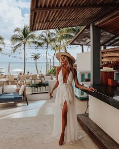 a woman in a white dress and straw hat standing on a patio next to the ocean