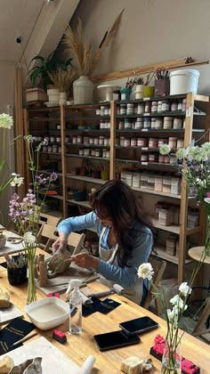 a woman working in a flower shop with flowers on the table and shelves behind her