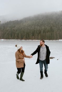 a man and woman holding hands walking in the snow near a lake with pine trees