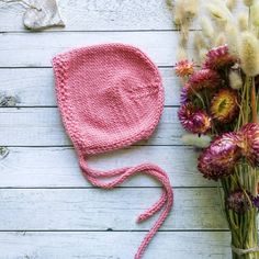 a pink knitted hat next to flowers on a white wooden surface with a string