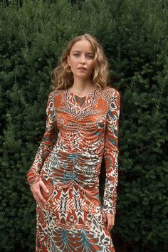 a woman standing in front of a bush wearing an orange and blue patterned long sleeved dress