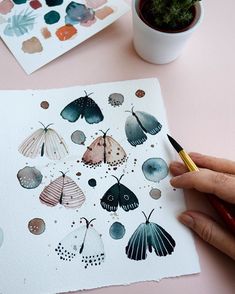 a person is holding a pen and drawing on paper with watercolors next to a potted plant