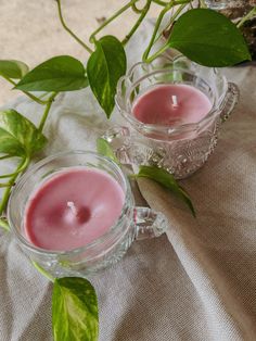 two tea cups filled with pink candles on top of a white table cloth next to green leaves