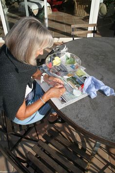 an older woman sitting at a table painting with watercolors on the paper she is holding