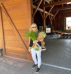 a woman standing in front of a wooden building holding a bag full of fresh vegetables