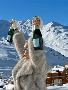 a woman holding two bottles of champagne in the snow with mountains in the back ground