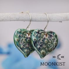 two green heart shaped ceramic earrings hanging from a twig on a white wooden stick