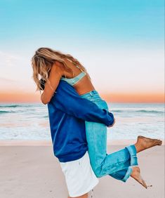 a woman in blue shirt and white pants on the beach with her arms around another woman's back