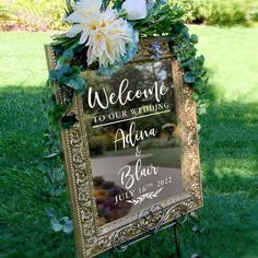 a wedding sign with flowers on it in the grass next to a mirror that says welcome to our wedding