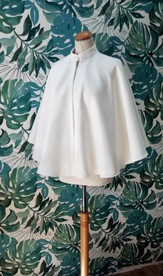 a white cape on a mannequin stand in front of a wall with green leaves