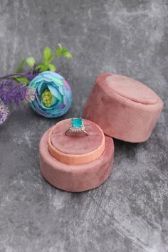 a pink velvet ring box with a turquoise stone in the center and flowers behind it