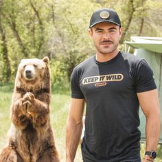 a man standing next to a brown bear in the grass with his hands on his hips