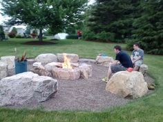 two men sitting around a fire pit in the middle of a yard with rocks and grass