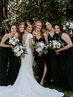 a group of women standing next to each other in front of trees and bushes holding bouquets