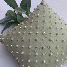 a green pillow with pearls on it sitting next to a leafy branch and some leaves